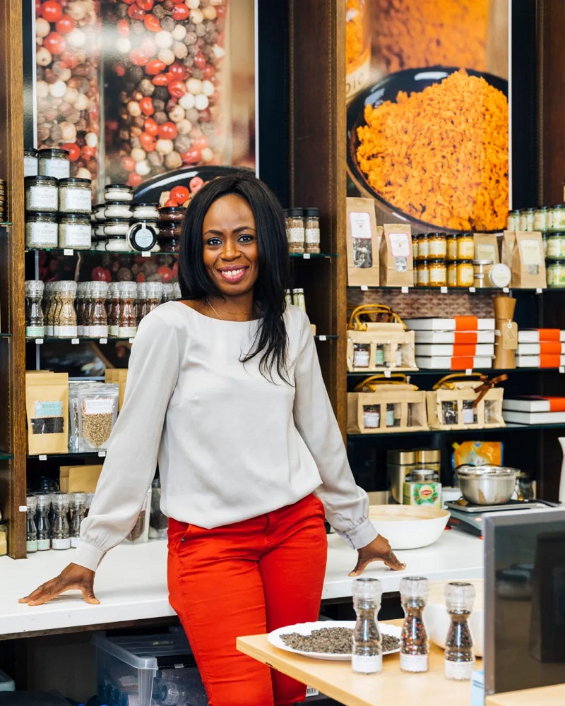 Cynthia Kouassi, fondatrice de STAR Collections, souriante dans sa boutique d’épices africaines, entourée de produits artisanaux et de moulins à poivre, avec des visuels colorés en arrière-plan.