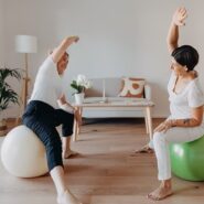 Deux femmes, dont l'une est identifiée comme Elise Bernold (à gauche), sont assises sur des ballons d'exercice (blanc et vert) avec leurs bras étirés au-dessus de la tête dans un exercice d'étirement latéral, dans une pièce lumineuse et moderne.