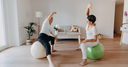 Deux femmes, dont l'une est identifiée comme Elise Bernold (à gauche), sont assises sur des ballons d'exercice (blanc et vert) avec leurs bras étirés au-dessus de la tête dans un exercice d'étirement latéral, dans une pièce lumineuse et moderne.