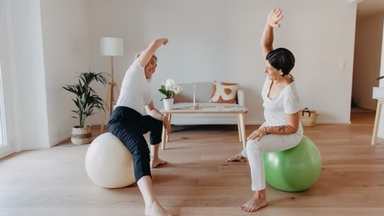 Deux femmes, dont l'une est identifiée comme Elise Bernold (à gauche), sont assises sur des ballons d'exercice (blanc et vert) avec leurs bras étirés au-dessus de la tête dans un exercice d'étirement latéral, dans une pièce lumineuse et moderne.