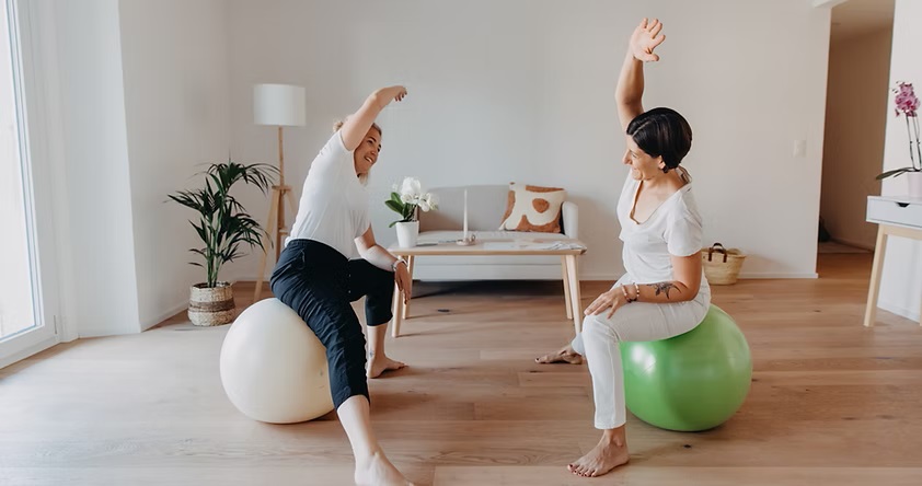 Deux femmes, dont l'une est identifiée comme Elise Bernold (à gauche), sont assises sur des ballons d'exercice (blanc et vert) avec leurs bras étirés au-dessus de la tête dans un exercice d'étirement latéral, dans une pièce lumineuse et moderne.