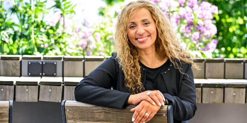 Leonilda Renaldo, assise à l’extérieur sur un banc en bois, souriante et détendue dans un cadre fleuri et lumineux.