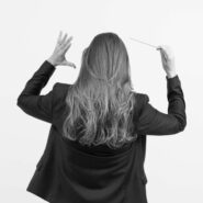 Lucie Leguay de dos, en train de diriger un orchestre, baguette à la main, vêtue d’une veste noire, sur fond blanc.