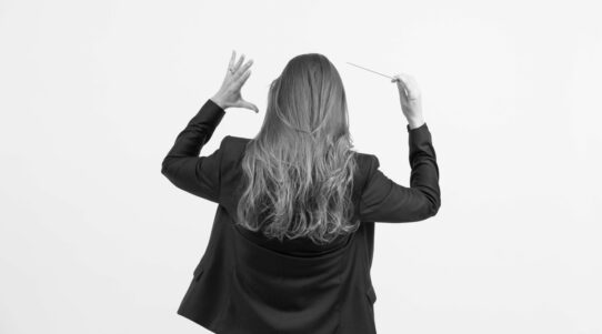 Lucie Leguay de dos, en train de diriger un orchestre, baguette à la main, vêtue d’une veste noire, sur fond blanc.