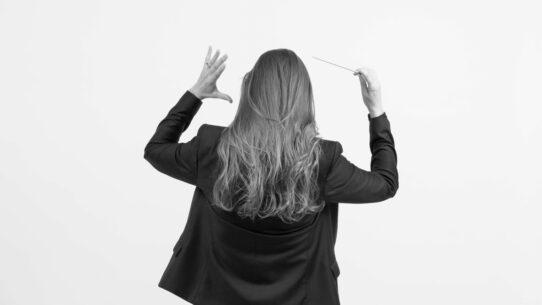 Lucie Leguay de dos, en train de diriger un orchestre, baguette à la main, vêtue d’une veste noire, sur fond blanc.