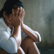 Une personne assise par terre, la tête dans les mains, semblant en détresse ou bouleversée, devant un mur faiblement éclairé.