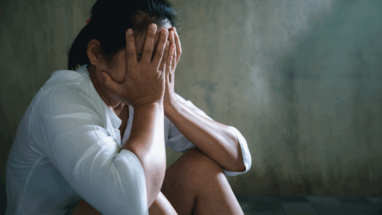 Une personne assise par terre, la tête dans les mains, semblant en détresse ou bouleversée, devant un mur faiblement éclairé.