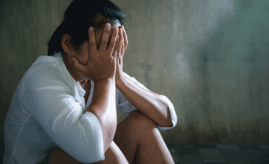 Une personne assise par terre, la tête dans les mains, semblant en détresse ou bouleversée, devant un mur faiblement éclairé.