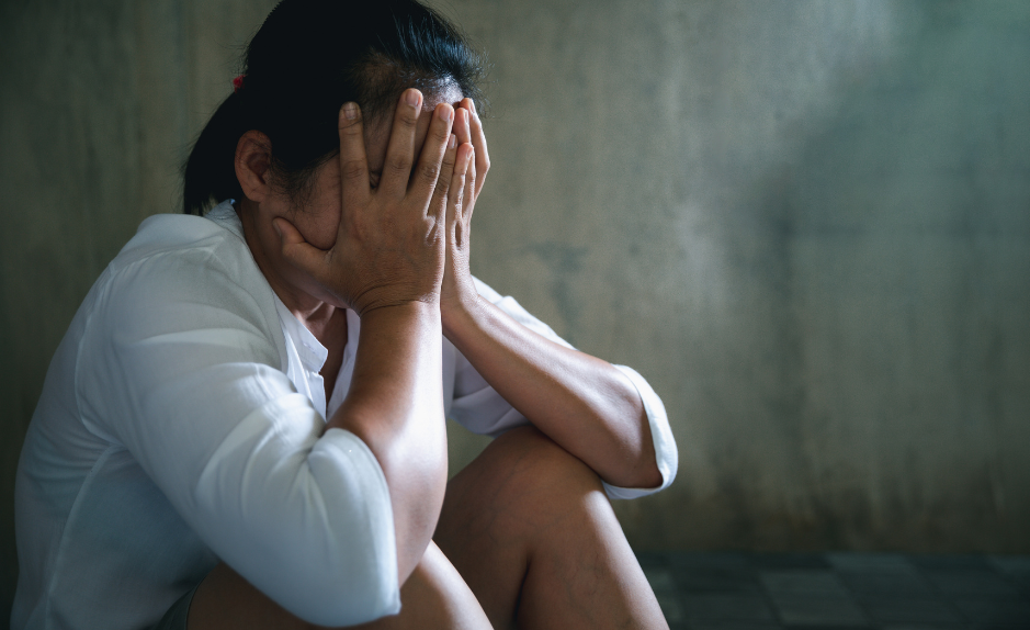 Une personne assise par terre, la tête dans les mains, semblant en détresse ou bouleversée, devant un mur faiblement éclairé.