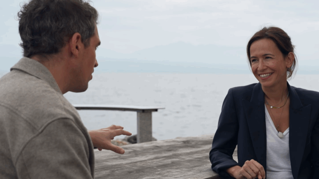 Lucia Rochat en discussion au bord de l’eau, souriante, assise à une table en bois avec une boîte de savon The Soap and the Sea — symbole de son engagement pour la protection des océans.