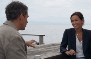 Lucia Rochat en discussion au bord de l’eau, souriante, assise à une table en bois avec une boîte de savon The Soap and the Sea — symbole de son engagement pour la protection des océans.