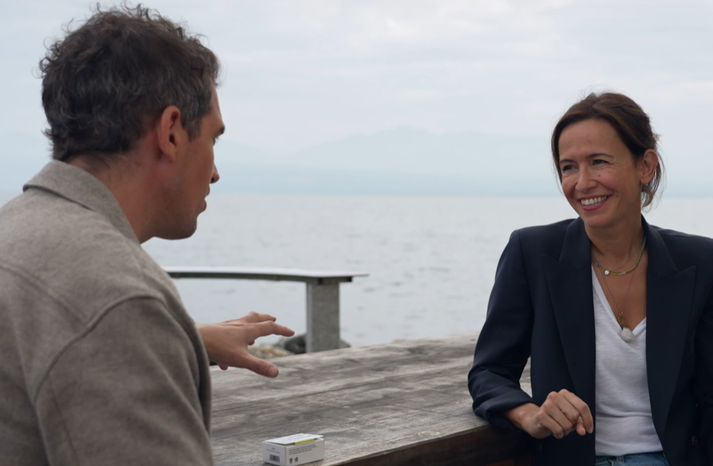 Lucia Rochat en discussion au bord de l’eau, souriante, assise à une table en bois avec une boîte de savon The Soap and the Sea — symbole de son engagement pour la protection des océans.
