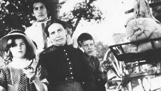 Photographie en noir et blanc d’Augusta Gillabert-Randin entourée de ses enfants, posant en plein air près d’une charrette agricole, au début du XXᵉ siècle.