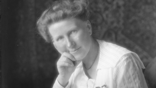 Portrait en noir et blanc d’Émilie Gourd, de trois-quarts, la tête légèrement inclinée et la main posée sous le menton, portant un chemisier clair. Elle regarde l’objectif avec une expression douce et réfléchie.