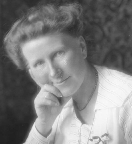 Portrait en noir et blanc d’Émilie Gourd, de trois-quarts, la tête légèrement inclinée et la main posée sous le menton, portant un chemisier clair. Elle regarde l’objectif avec une expression douce et réfléchie.