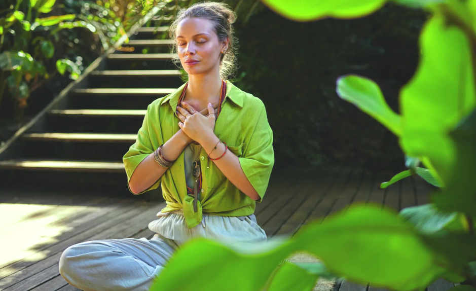 Femme assise en pleine nature, les yeux fermés et les mains posées sur la poitrine, illustrant un moment de respiration consciente et de régulation émotionnelle.