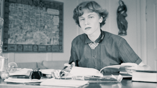 Iris von Roten assise à son bureau, écrivant et consultant des documents, dans un intérieur sobre, photographie en noir et blanc