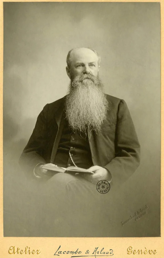 Portrait en sépia de Jean-Jacques Gourd assis, âgé, avec une longue barbe blanche, tenant un livre ouvert sur ses genoux, photographié par l’atelier Lacombe & Arlaud à Genève.