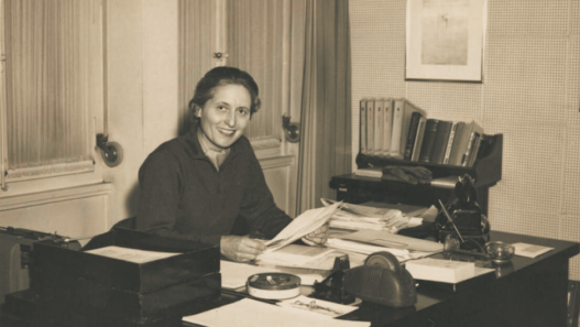 Marthe Gosteli assise à son bureau, souriante, entourée de dossiers et de documents, illustrant son travail d’archiviste et de militante pour la mémoire des femmes suisses.