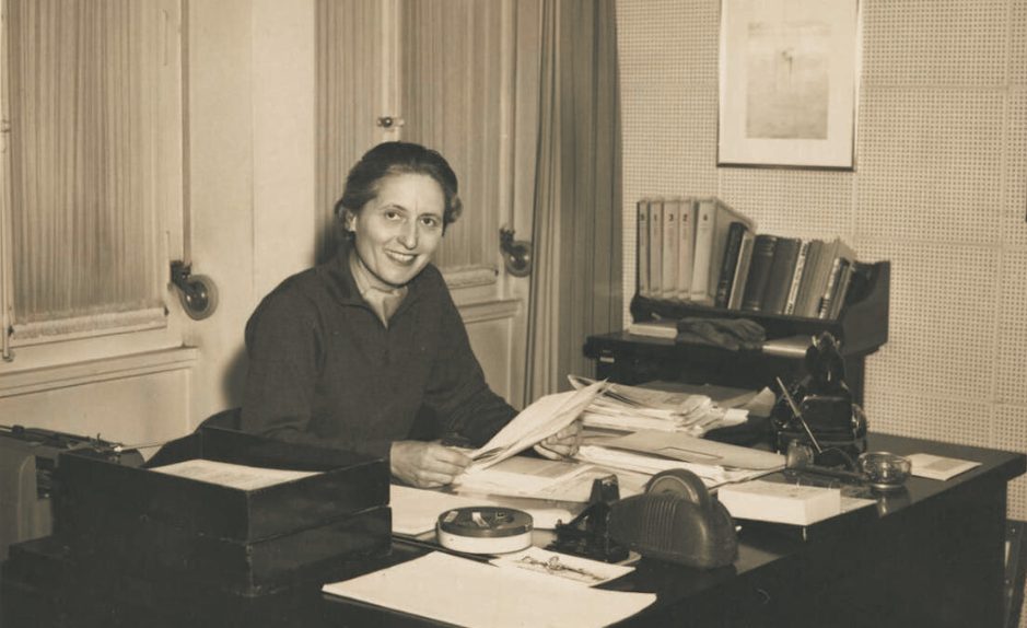 Marthe Gosteli assise à son bureau, souriante, entourée de dossiers et de documents, illustrant son travail d’archiviste et de militante pour la mémoire des femmes suisses.