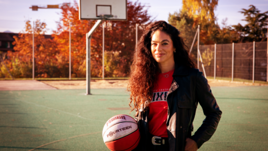 Portrait de Mouna Skaria tenant un ballon de basket sur un terrain extérieur, avec un panneau de basket en arrière-plan.