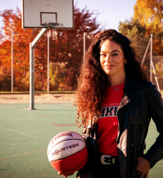 Portrait de Mouna Skaria tenant un ballon de basket sur un terrain extérieur, avec un panneau de basket en arrière-plan.