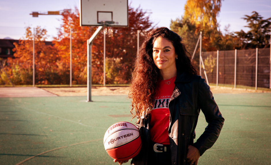 Portrait de Mouna Skaria tenant un ballon de basket sur un terrain extérieur, avec un panneau de basket en arrière-plan.