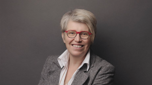 Portrait de Rébecca Dougoud, future directrice des Chemins de fer du Jura, souriante, portant des lunettes rouges et un blazer gris sur fond sombre.