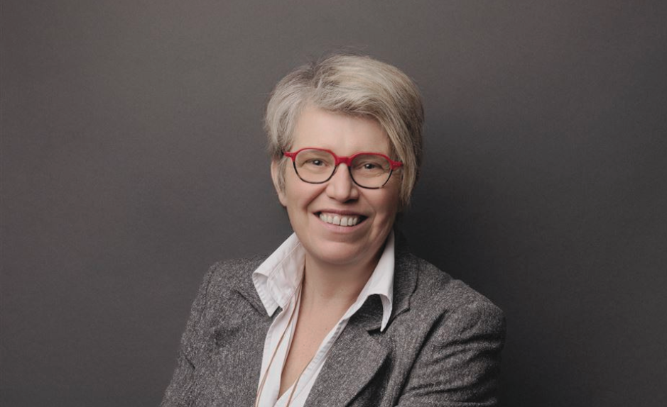 Portrait de Rébecca Dougoud, future directrice des Chemins de fer du Jura, souriante, portant des lunettes rouges et un blazer gris sur fond sombre.