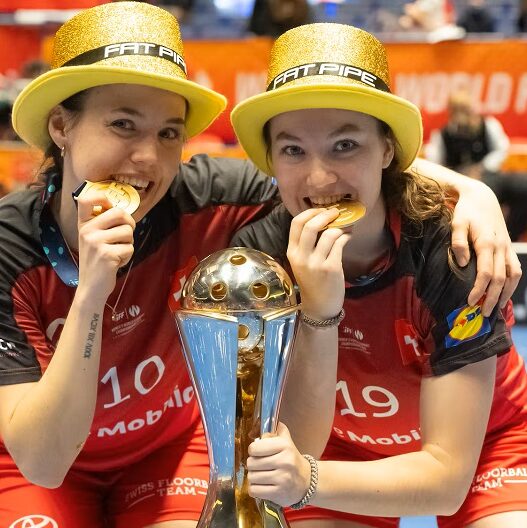 Deux joueuses de l’équipe de Suisse féminine de unihockey célèbrent leur titre de championnes du monde en croquant leur médaille d’or, trophée à la main.