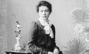 Portrait en noir et blanc de Clara Ragaz assise, regardant l’objectif, portant une robe sombre à manches longues et un chemisier clair à col montant noué, les cheveux relevés, devant un fond neutre avec un vase de fleurs posé à côté.