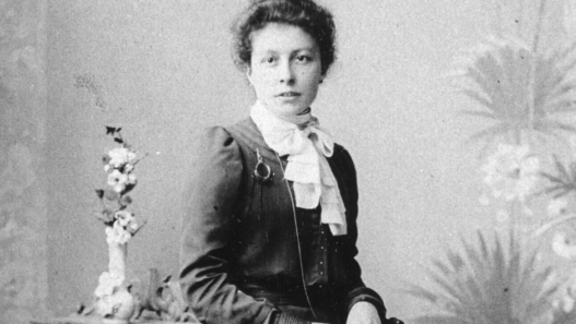 Portrait en noir et blanc de Clara Ragaz assise, regardant l’objectif, portant une robe sombre à manches longues et un chemisier clair à col montant noué, les cheveux relevés, devant un fond neutre avec un vase de fleurs posé à côté.