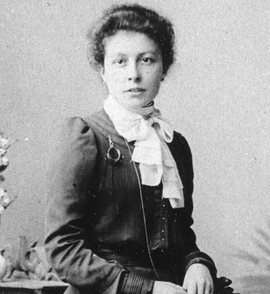 Portrait en noir et blanc de Clara Ragaz assise, regardant l’objectif, portant une robe sombre à manches longues et un chemisier clair à col montant noué, les cheveux relevés, devant un fond neutre avec un vase de fleurs posé à côté.