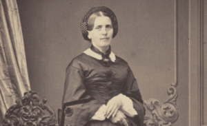 Photographie ancienne en sépia montrant Johanna Spyri, les mains croisées, posant devant un décor intérieur avec rideau et dossier de chaise sculpté.