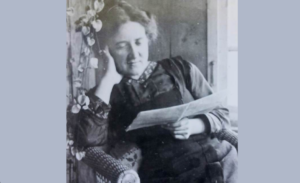 Portrait en noir et blanc de Marguerite Gobat dans un fauteuil, la tête appuyée sur une main, lisant une feuille de papier près d’une fenêtre.