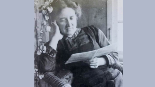 Portrait en noir et blanc de Marguerite Gobat dans un fauteuil, la tête appuyée sur une main, lisant une feuille de papier près d’une fenêtre.