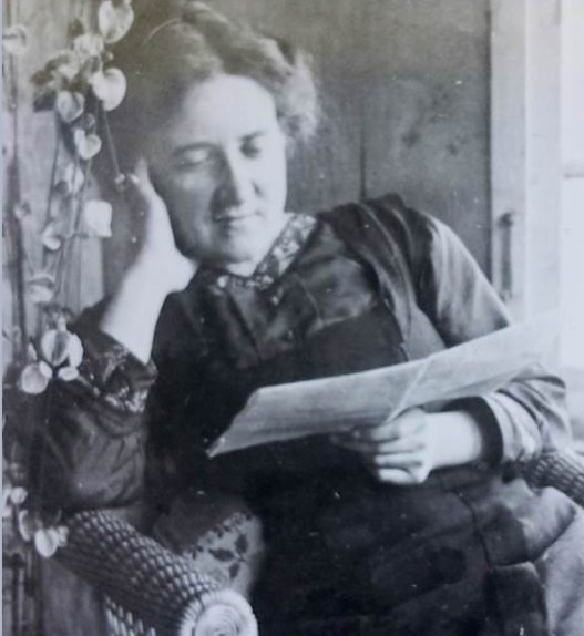 Portrait en noir et blanc de Marguerite Gobat dans un fauteuil, la tête appuyée sur une main, lisant une feuille de papier près d’une fenêtre.