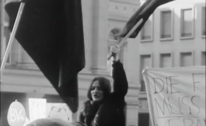 Manifestation féministe en Suisse devant un bâtiment officiel, une femme brandit un drapeau lors d’un rassemblement pour les droits des femmes.