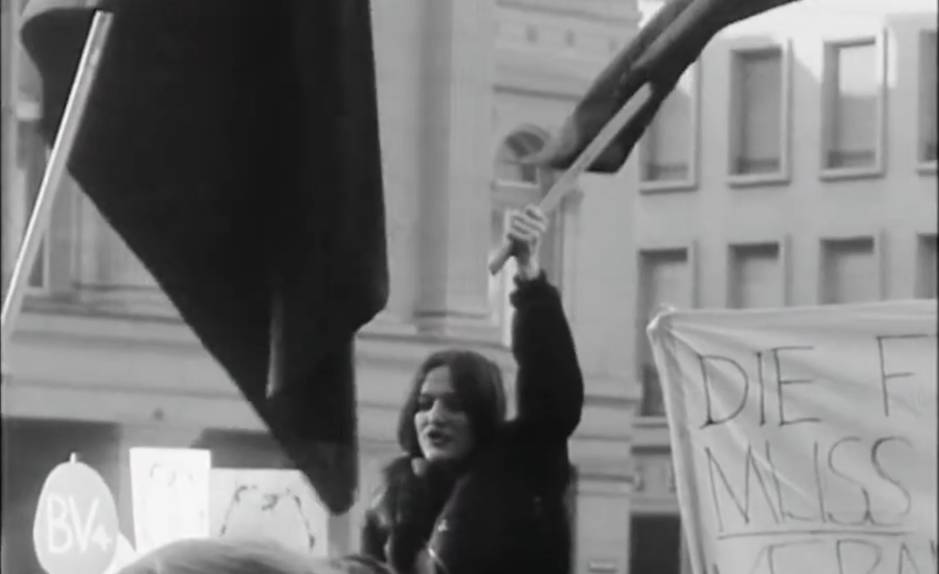 Manifestation féministe en Suisse devant un bâtiment officiel, une femme brandit un drapeau lors d’un rassemblement pour les droits des femmes.