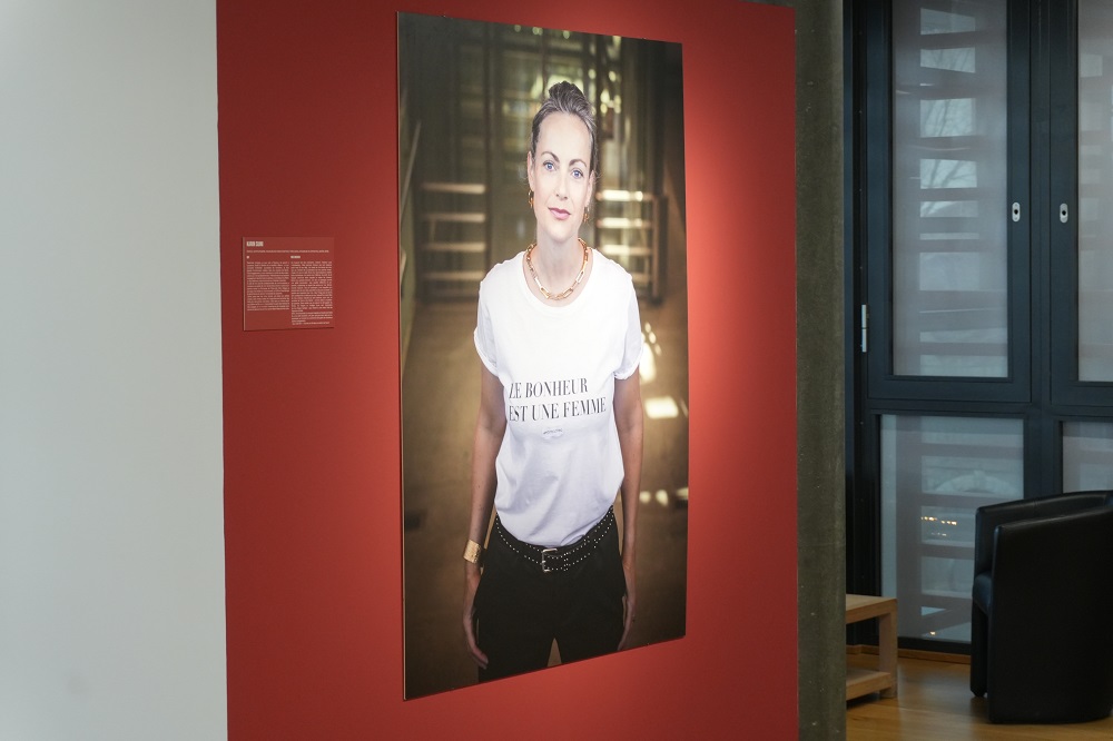 Portrait photographique d’une femme aux cheveux gris attachés, exposé à la Galerie du Boléro à Versoix dans le cadre de l’exposition Silver Power. Elle porte un t-shirt blanc avec le message “Le bonheur est une femme”.
