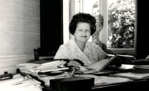Portrait en noir et blanc de Lise Girardin assise à son bureau, entourée de dossiers et de documents, dans un bureau lumineux avec une fenêtre en arrière-plan.