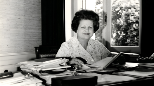 Portrait en noir et blanc de Lise Girardin assise à son bureau, entourée de dossiers et de documents, dans un bureau lumineux avec une fenêtre en arrière-plan.