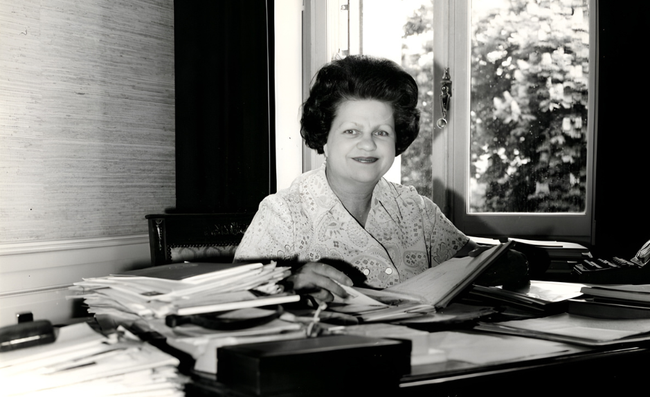 Portrait en noir et blanc de Lise Girardin assise à son bureau, entourée de dossiers et de documents, dans un bureau lumineux avec une fenêtre en arrière-plan.