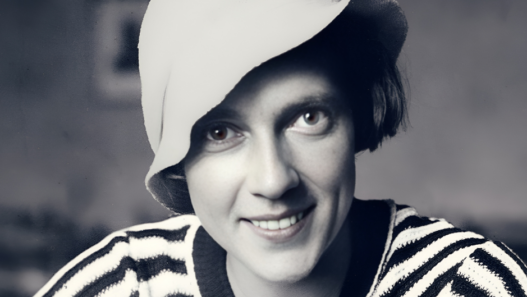 Portrait en noir et blanc d’Ella Maillart jeune, souriante, portant un chapeau clair et un pull rayé