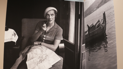 Photographie en noir et blanc d’une voyageuse assise dans un compartiment de train, une pipe à la main, une grande carte dépliée sur les genoux ; à droite, image d’un bateau sur l’eau, extrait de l’exposition « Ella Maillart. Récits photographiques » à Photo Elysée (Lausanne).