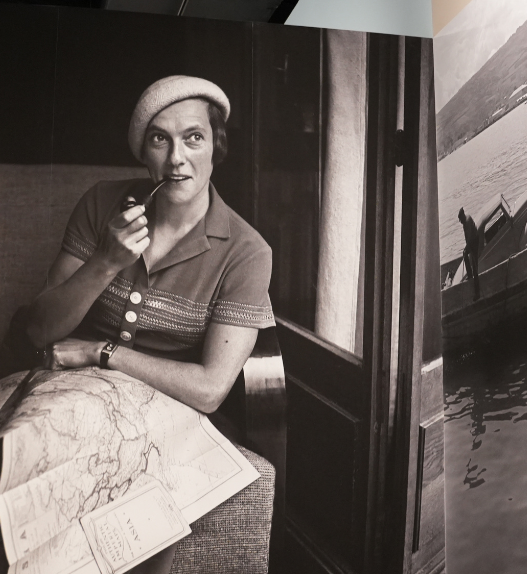Photographie en noir et blanc d’une voyageuse assise dans un compartiment de train, une pipe à la main, une grande carte dépliée sur les genoux ; à droite, image d’un bateau sur l’eau, extrait de l’exposition « Ella Maillart. Récits photographiques » à Photo Elysée (Lausanne).