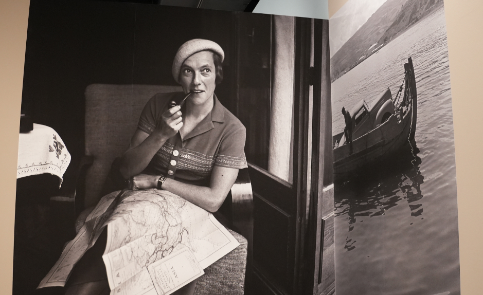 Photographie en noir et blanc d’une voyageuse assise dans un compartiment de train, une pipe à la main, une grande carte dépliée sur les genoux ; à droite, image d’un bateau sur l’eau, extrait de l’exposition « Ella Maillart. Récits photographiques » à Photo Elysée (Lausanne).