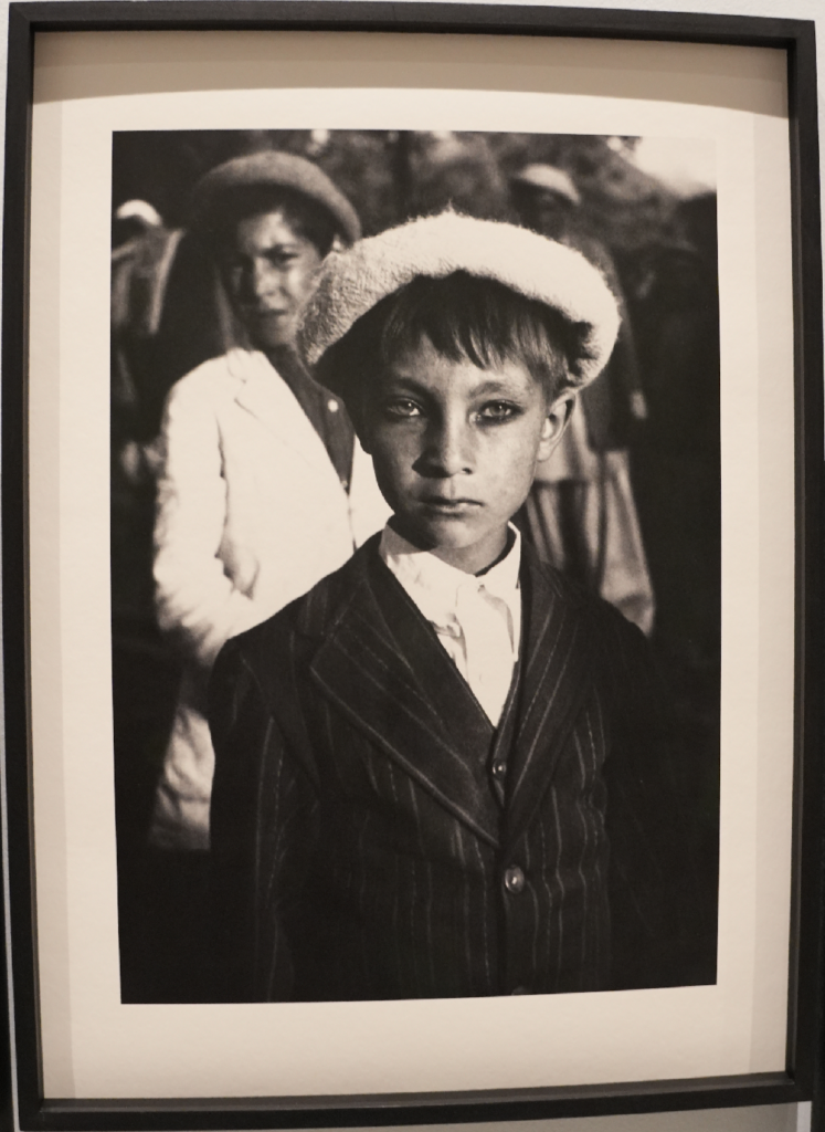 Photographie en noir et blanc encadrée : portrait d’un garçon portant un béret et un costume rayé, regard face caméra, avec un groupe flou en arrière-plan.