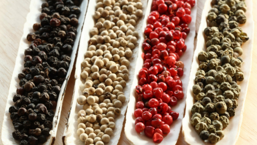 Quatre rangées de grains de poivre noir, blanc, rouge et vert alignées côte à côte sur des supports clairs, montrant visuellement leurs différences de couleur et d’aspect.