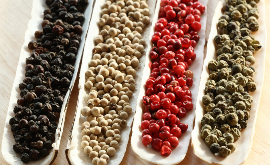 Quatre rangées de grains de poivre noir, blanc, rouge et vert alignées côte à côte sur des supports clairs, montrant visuellement leurs différences de couleur et d’aspect.